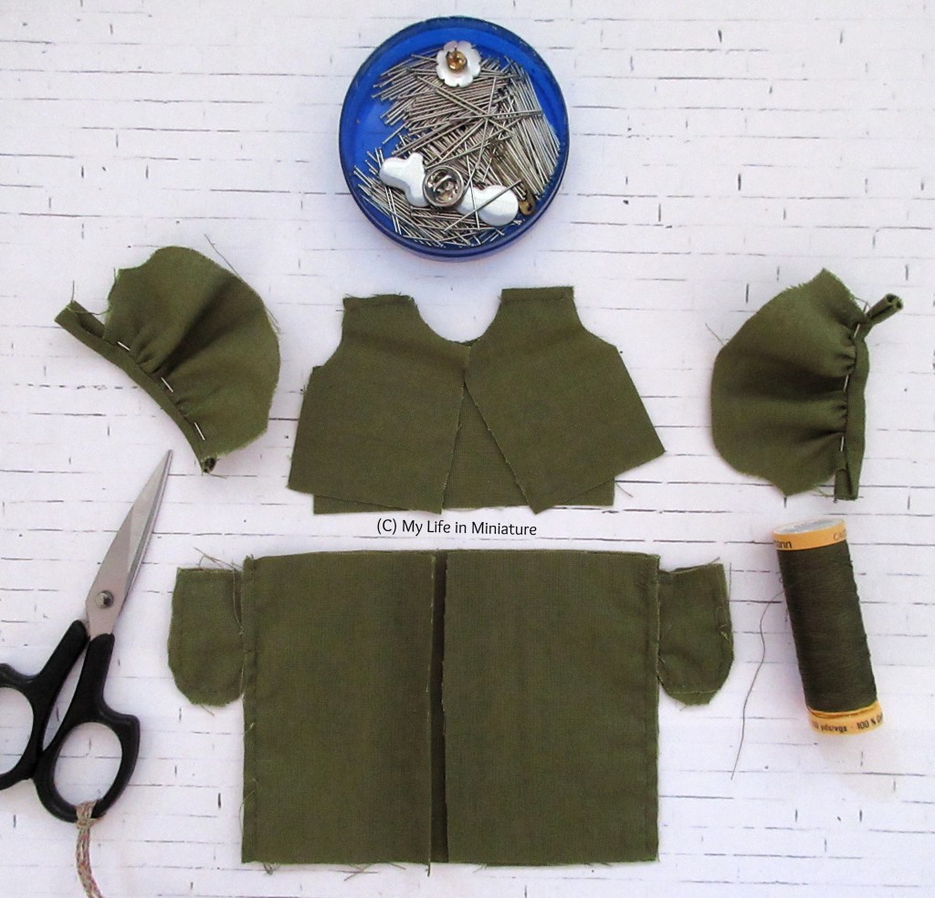 The pieces of a dress, partially assembled, are laid out on a white brick background. All the pieces are khaki green, the skirt below the bodice pieces in the centre and the sleeves beside the bodice. Around the dress is a pin tin, a reel of khaki thread, and a pair of black embroidery scissors. 