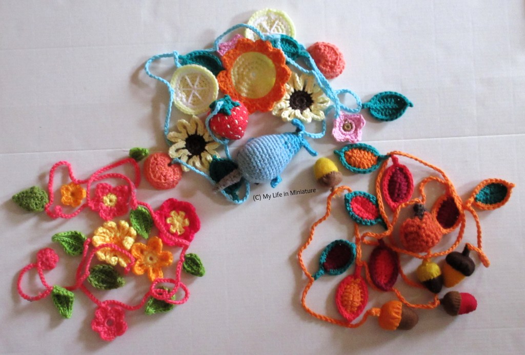 The summer garland is coiled in a heap on a white background, along with a spring and an autumn garland, coiled in similar heaps. The spring garland is quite pink and floral, and the autumn garland is very orange and leafy.