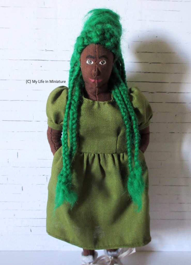 Hazel stands in front of a white brick background, two green braids pushed forward over each shoulder. Her hands are in the pockets of her khaki green dress, which is midi-length and has short, puffy sleeves. She wears white lace-up shoes and smiles at the camera.