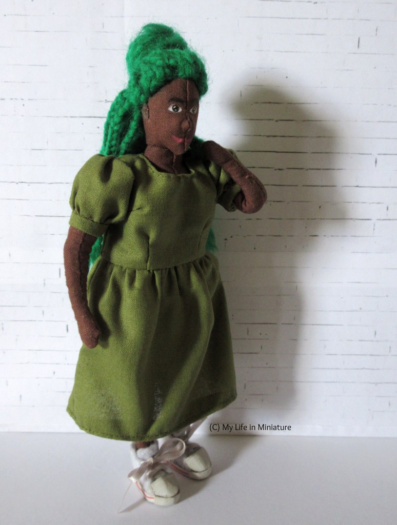 Hazel stands in front of a white brick background. She wears a midi-length khaki green dress with puffed sleeves and white shoes. She;s looking to the right of the camera with one hand behind her neck and the other at her side.