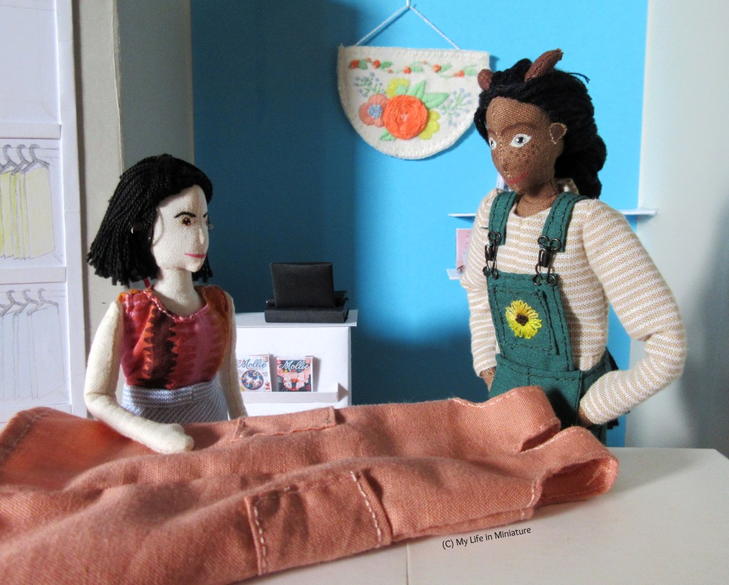 Petra and Tiffany stand at one of Needle & Thread's cutting tables. A peach pink dress is laid out on the table. Tiffany is smoothing the fabric, and Petra looks at it, excited. Tiffany wears a multi-coloured top with a blue striped skirt. Petra wears green dungarees with a beige striped long-sleeved shirt.