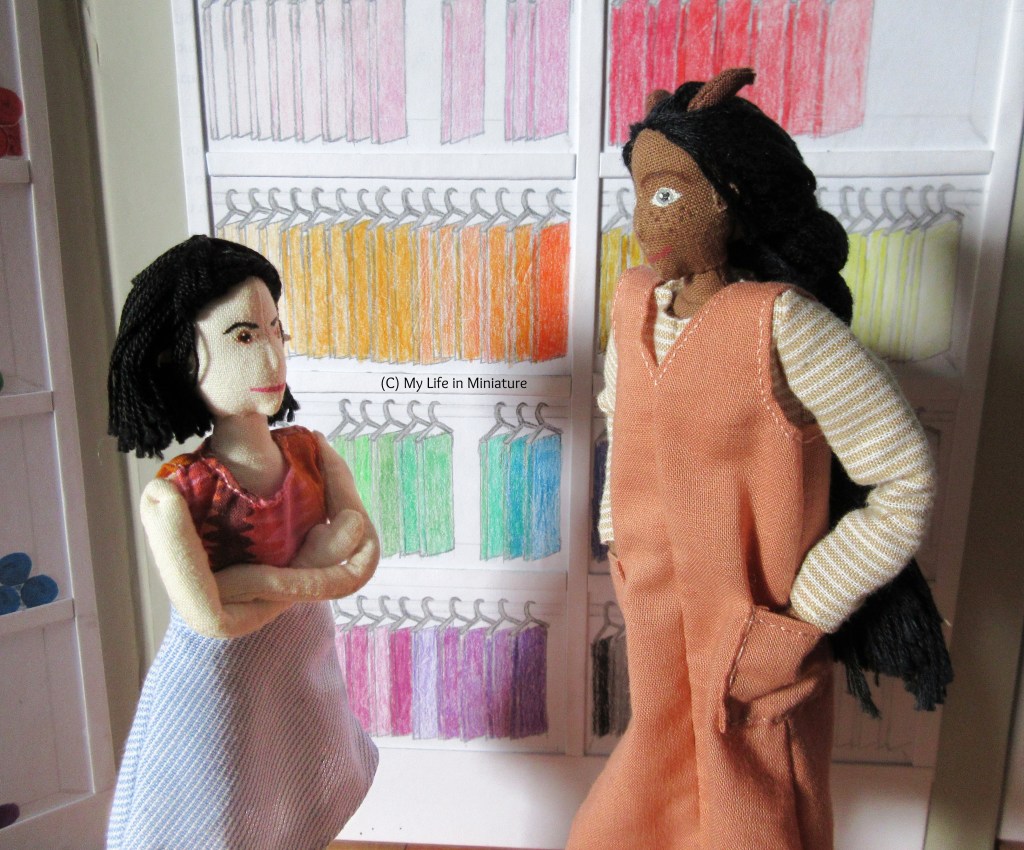 Petra and Tiffany stand side by side in front of Needle & Thread's fat quarters. They are looking at each other as Petra thanks Tiffany for the dress. Petra's hands are still in the pockets of the dress, while Tiff's are still crossed. 