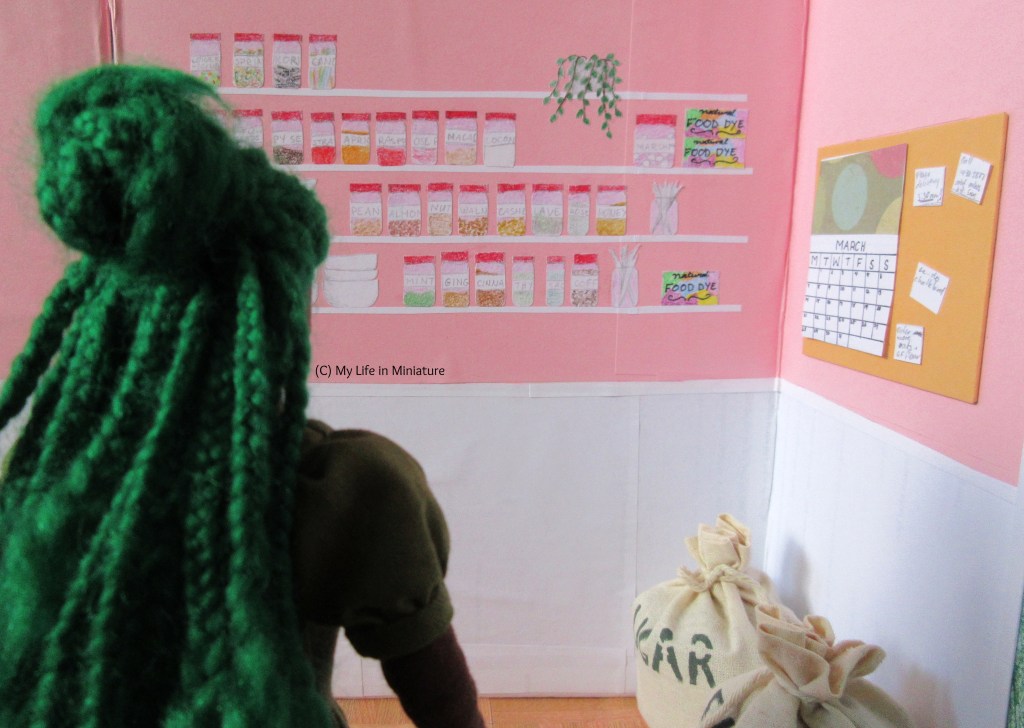 Hazel stands in the foreground, her back to the camera. She looks at the four white shelves on the opposite wall of Fierro's back room, which are now full. (More jars of ingredients and boxes of food dye have been added to the top two shelves.) On the wall to Hazel's right there is an orange noticeboard above the sugar and flour sacks. The noticeboard has a calendar and several reminders pinned to it.