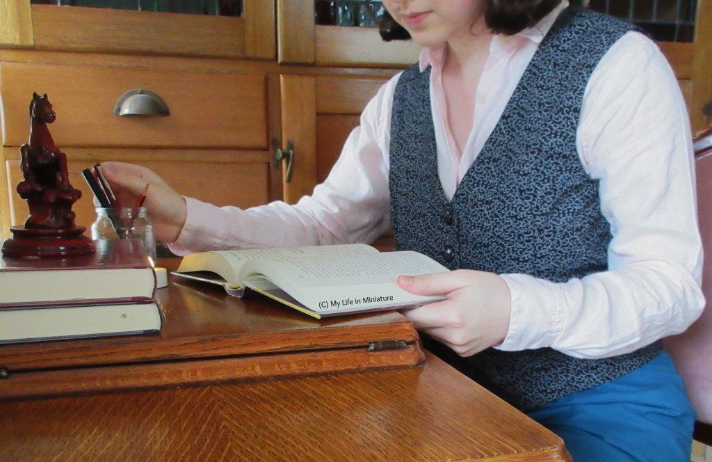 The author sits at an antique wooden-topped desk, with a wooden cabinet behind her. She wears a black waistcoat patterned with grey swirls over a pale pink long-sleeves shirt with turquoise pants. She holds a hardback book open as she reaches for one of the pens in the pen jar. 
