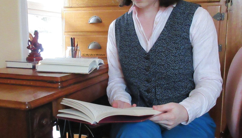The author sits at an antique wooden-topped desk, with a wooden cabinet behind her. She wears a black waistcoat patterned with grey swirls over a pale pink long-sleeves shirt with turquoise pants. She is turned sideways in the upholstered wooden chair she sits in, with another book open on her lap.