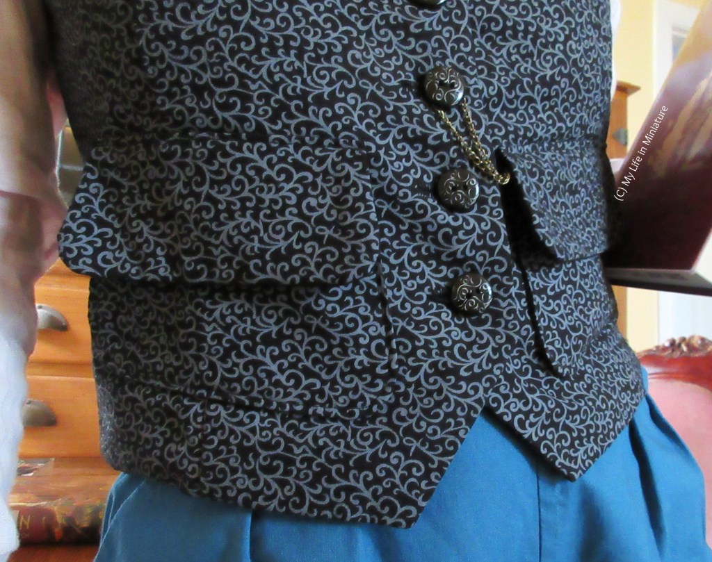 Close-up of the bottom half of the black and grey waistcoat. The bottom three buttons are visible, as well as both of the pockets. There is also a brass watch chain looped around one of the buttons that disappears into one of the pockets. 