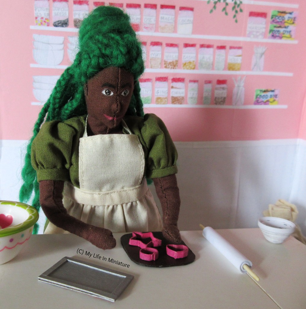 Hazel stands at the work bench in Fierro's. All the cutters have been positioned on the rolled-out dough, and Hazel's hand hovers above the heart-shaped one, about to press it into the dough.