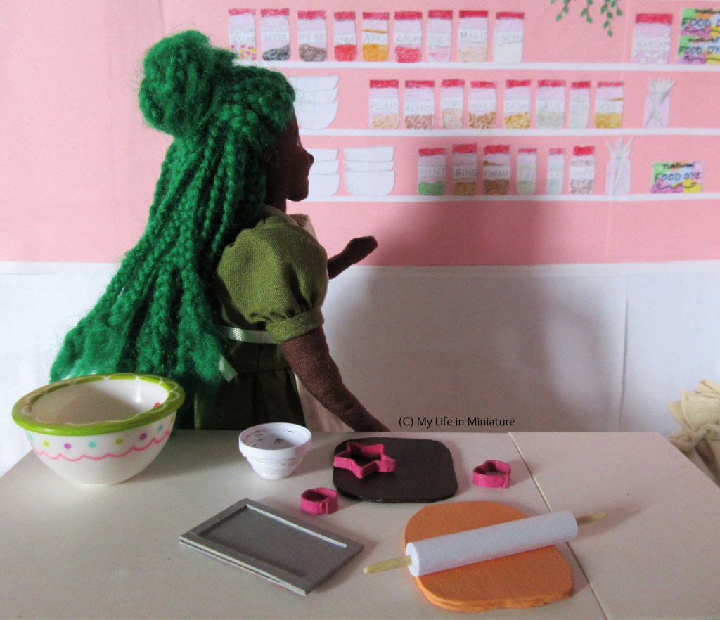 Hazel is turned towards the wall of decorations and tools behind her, one arm raised as she searches for something. The big mixing bowl, flour bowl, two doughs, cutters, baking tray, and rolling pin are all still on the bench, as if the baker jumped to a new idea before finishing here. 