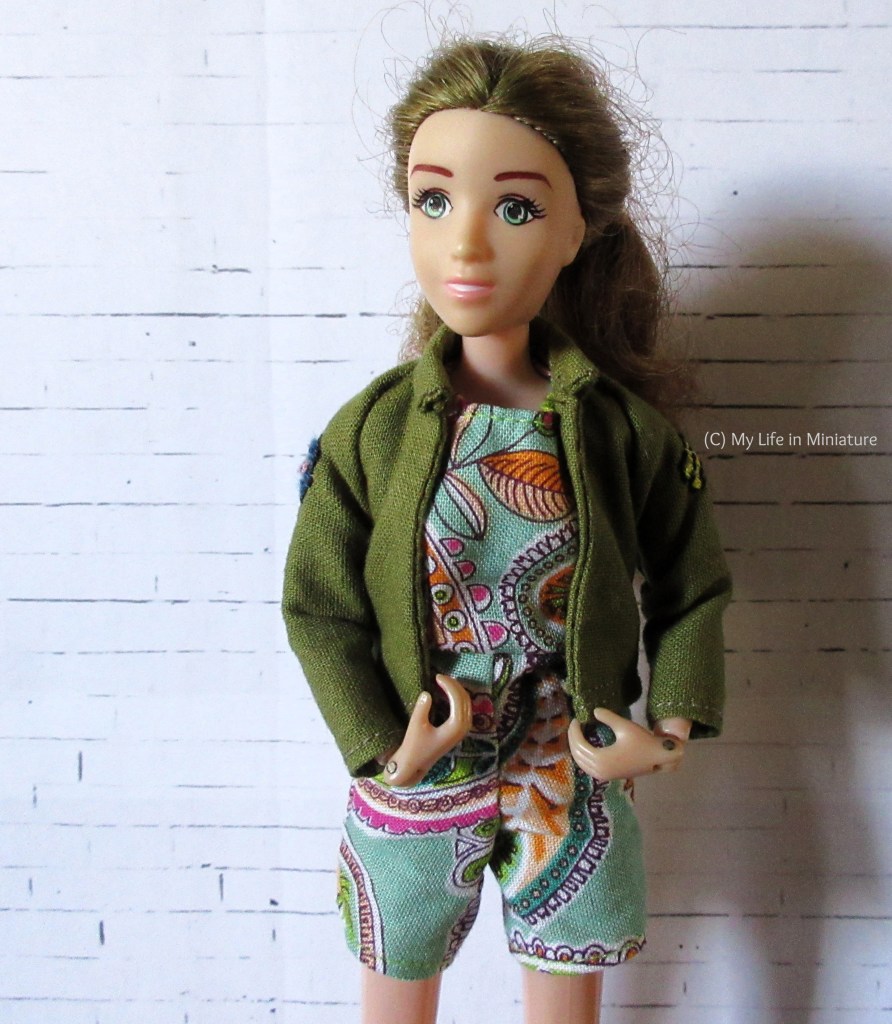 Sarah, a white-skinned woman with brownish hair, stands in front of a white brick background. She wears a turquoise playsuit with paisley patterns underneath a khaki jacket that has embroidery on the sleeves. The jacket is clearly too small, and has lots of wrinkles at the shoulder-underarm area. Sarah has a hand on each front panel of the jacket, and looks to the left of the camera.