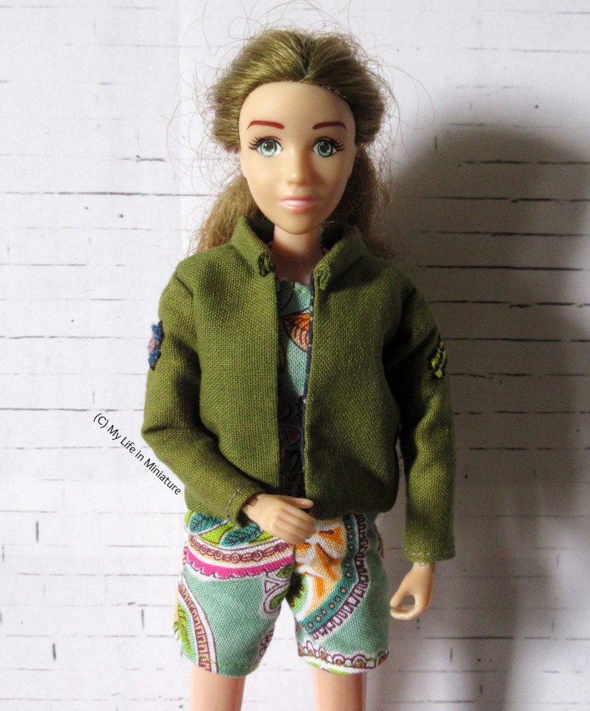 Sarah looks straight at the camera, smiling. She wears the turquoise playsuit underneath a very similar khaki jacket. The sleeves have the same embroidery on them, and the collar is the same stand-up style, but the torso fits her much better. A white brick background is behind her.