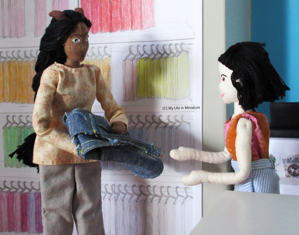 Petra, a tall brown woman, and Tiffany, a petite Asian woman, stand in Needle & Thread. Petra looks at Tiffany, holding a folded-up denim garment in her arms. She wears a cream long-sleeved shirt with grey pants. Tiffany has her arms out to receive the denim garment. She wears a pink and orange sleeveless shirt and a blue striped skirt. Behind Petra are racks of multi-coloured fat quarters, and in front of Tiffany is the end of a cutting table.