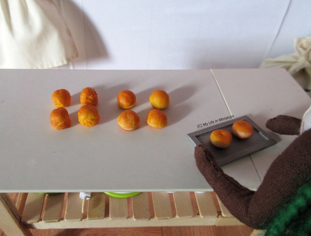 Hazel is in the back room of Fierro's bakery, and she is putting a small baking tray onto the main work surface. She is in the bottom left of the image, with her dark brown arms and hands visible. Also sitting on the table are four scones and four bread rolls, nicely browned from the oven. 