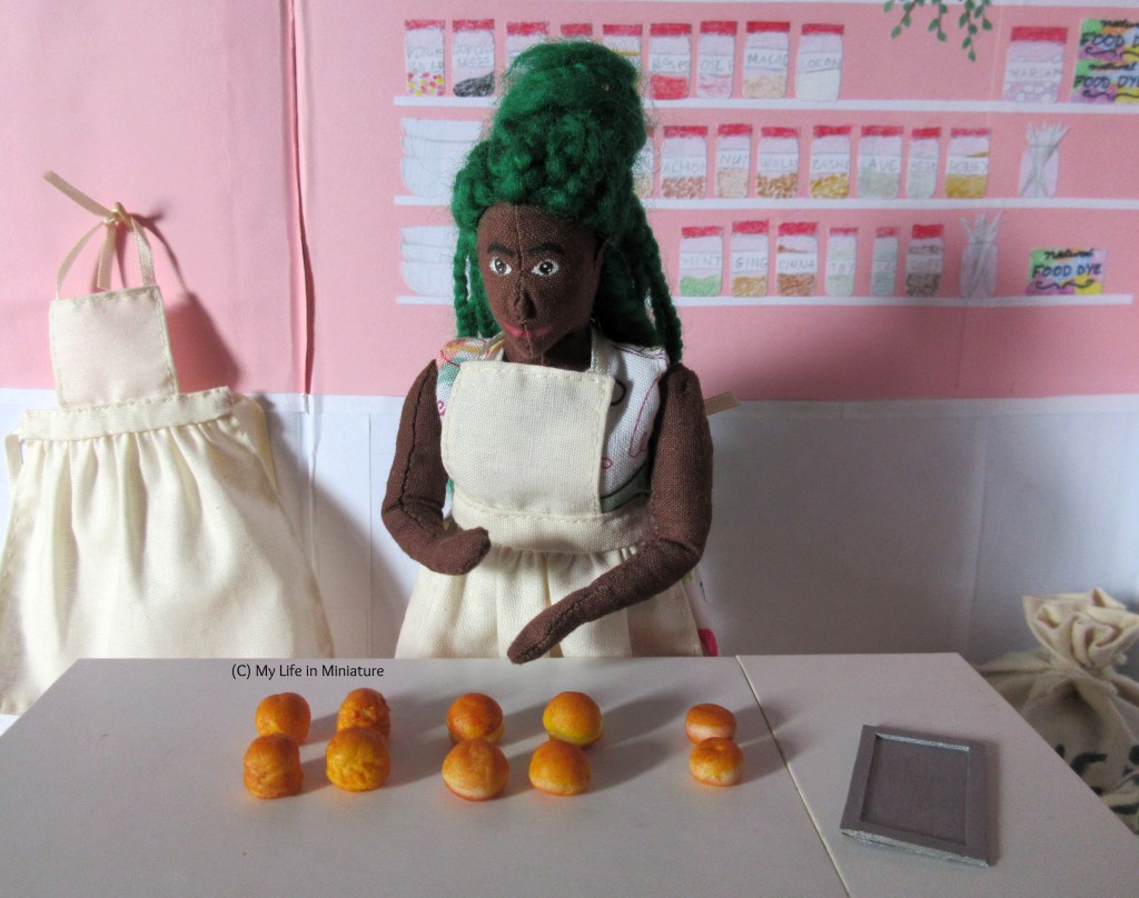 Hazel stands behind the main work bench in Fierro's, brushing excess flour off her hands. She wears an apron over her floral dress, and her green braids are in a half-up bun. On the work bench in front of her are four scones, four bread rolls, two small bagels, and a small baking tray. Behind Hazel and to the left of frame is a wooden hook, from which hangs another apron.