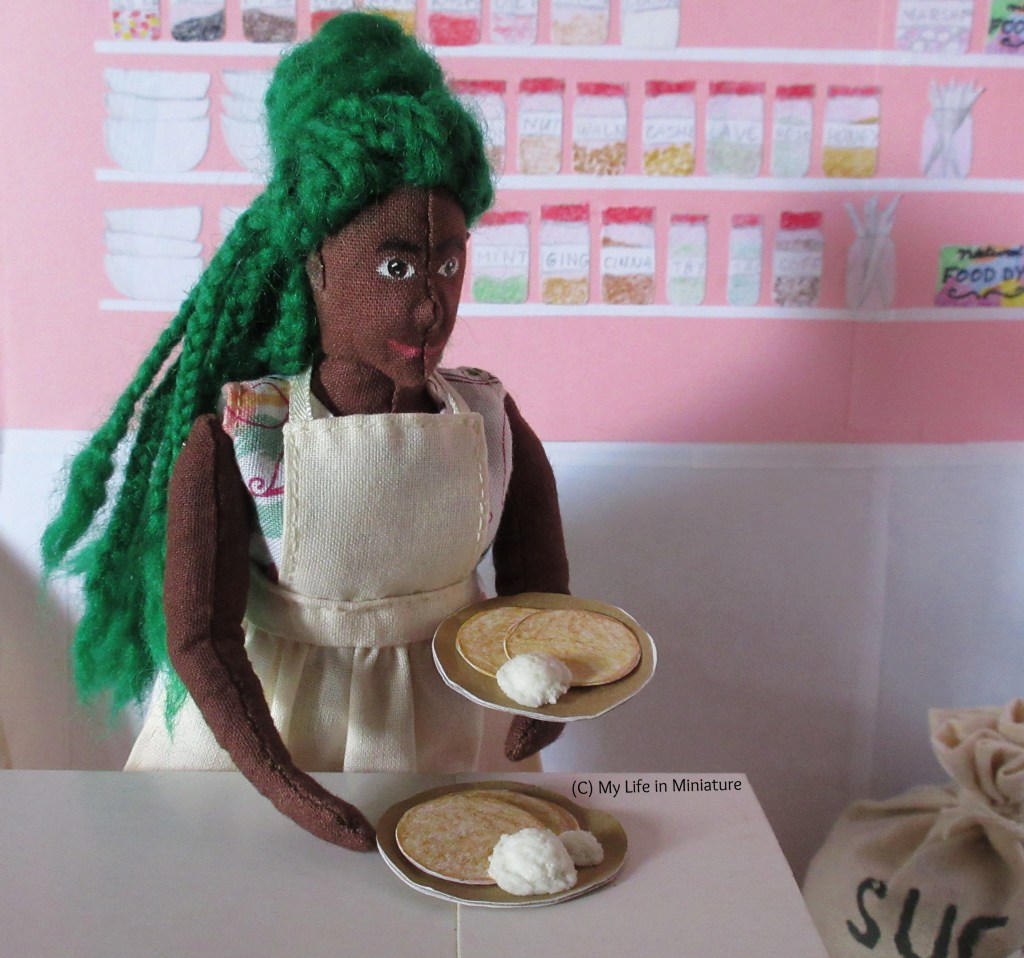 Hazel is in the back room of Fierro's, wearing the same apron-over-floral-dress outfit as before. She moves towards the doorway, holding a plate with two pancakes and a scoop of vanilla ice cream on. As she moves, she reaches to grab another plate of pancakes and ice cream with her other hand.