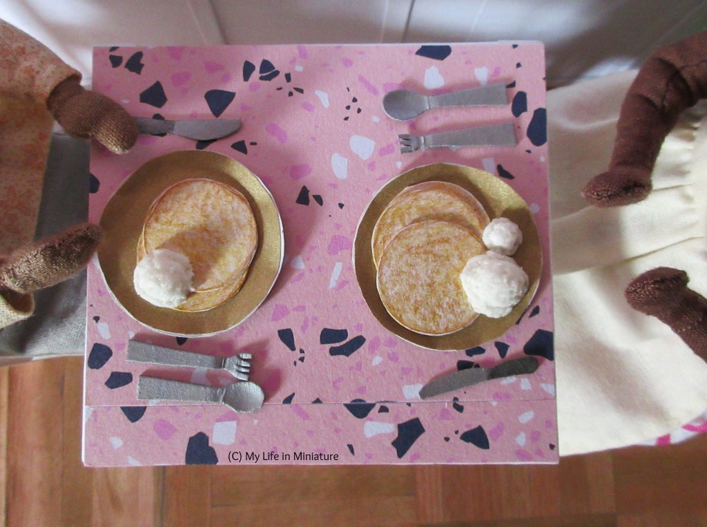 Overhead shot of the table Hazel and Petra sit at. The pancakes and ice cream are easier to see now, as they sit on gold plates. you can also see the cutlery well, and the pink terrazzo tabletop. Hazel and Petra's hands clap excitedly and move towards the cutlery, respectively.