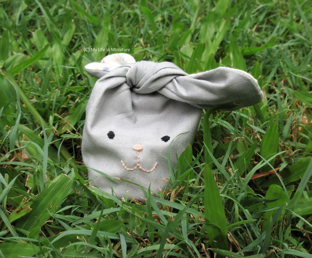 A small fabric bag sits outside, on grass. It is made to look like a rabbit, with an embroidered face and two 'ears' tied together to close the bag. It is pale grey, and the inside of the ears are made of white fabric, printed with blue flowers.