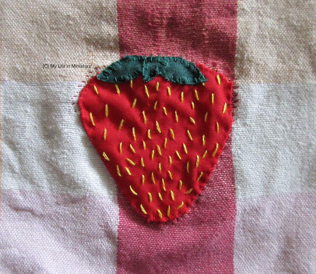 Close-up of the completed strawberry patch. There are now dark green leaves on the top of the strawberry, and the entirety of the red fabric is covered in yellow seed stitches. 