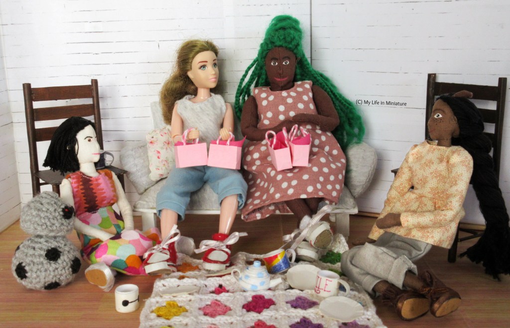 Tiffany, Sarah, Hazel, and Petra sit around a crocheted blanket spread on Sarah's living room floor. Tiffany and Petra sit on the left and right of the shot respectively, cross-legged and leaning against dining chairs. Chip the android is beside Tiffany. Sarah and Hazel sit on Sarah's couch, with the blanket at their feet. They each have two pink Fierro's gift bags on their laps, with pink tissue paper poking out of the top. On the blanket are four plates, three mugs, and a teapot. 