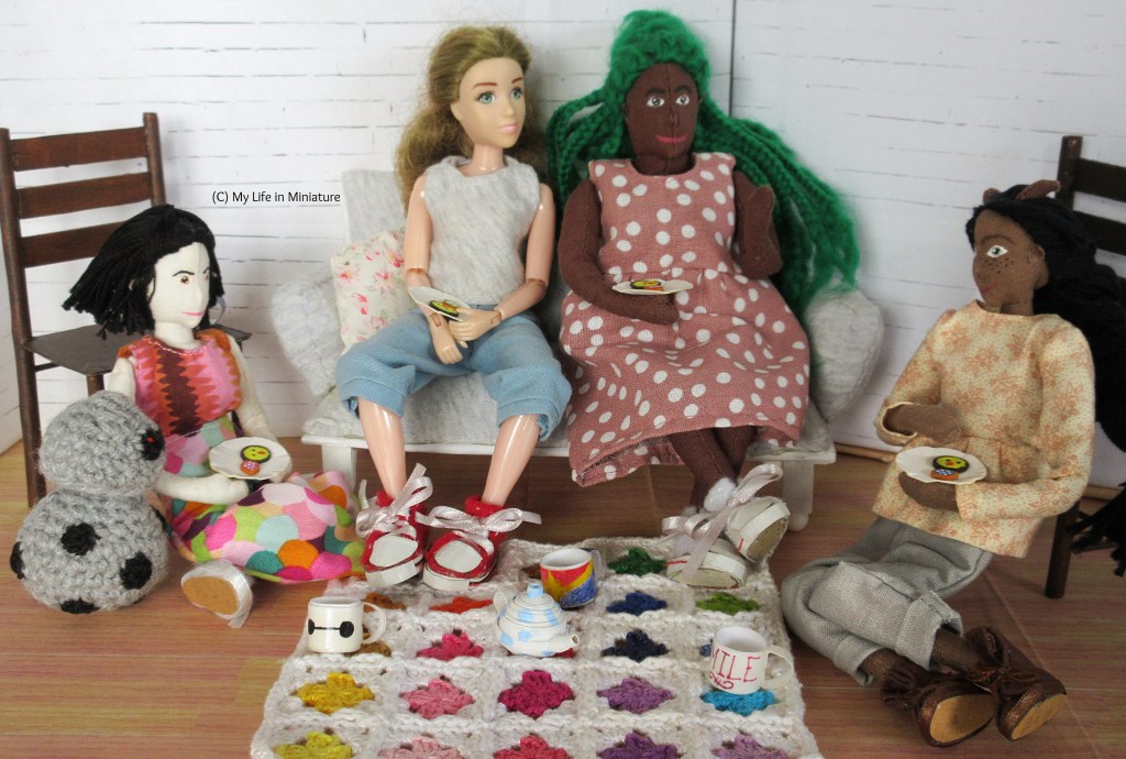Tiffany, Sarah, Hazel, and Petra sit around a crocheted blanket spread on Sarah's living room floor. They each hold a plate with the two biscuits on. Petra is picking up one of her biscuits as she listens to Hazel talk. Hazel is gesturing as she talks. Sarah and Tiffany listen on. Chip is looking closely at Tiffany's biscuits over her arm.