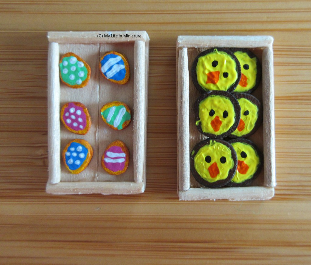 Close-up of two of Fierro's wooden display case trays. One is filled with six of the egg shaped biscuits, and the other with six chick-faced biscuits. There are three spotty egg biscuits and three stripy ones, one of each pattern in each colour.