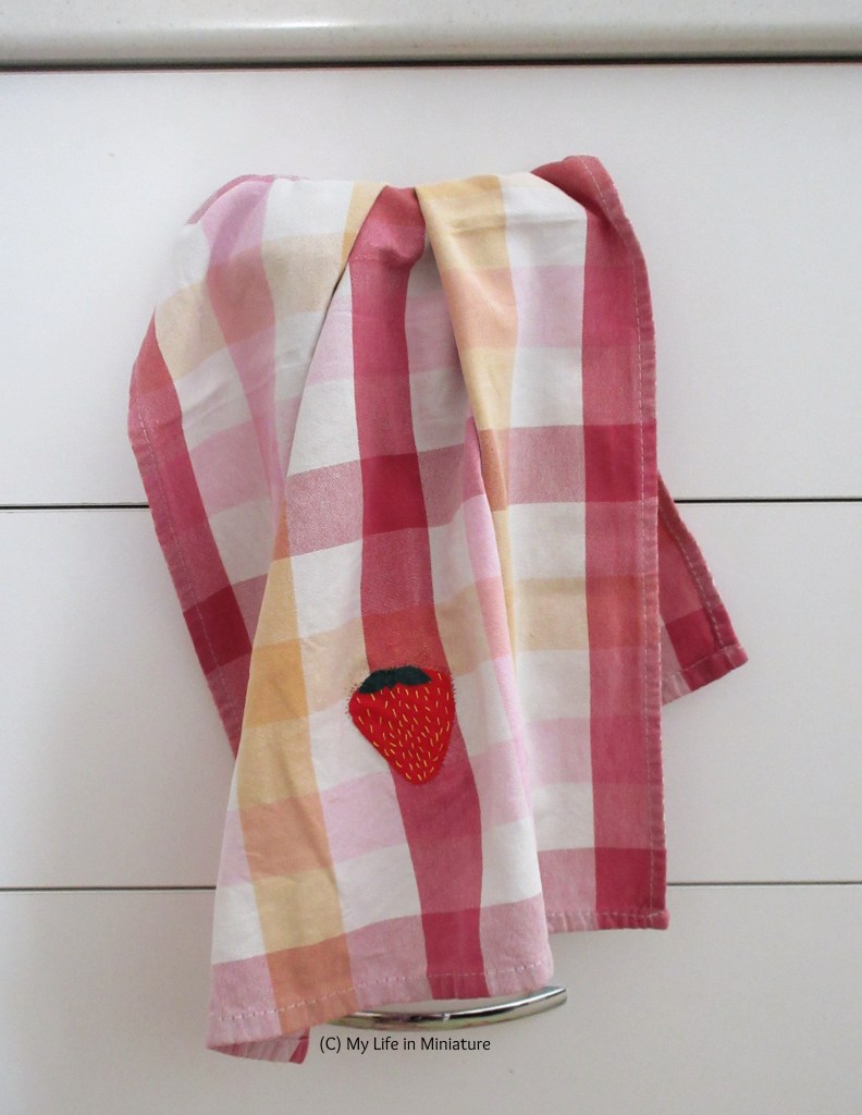 The pink checked tea towel hangs from the handle of a drawer in a kitchen. The completed strawberry patch is visible on the front. 