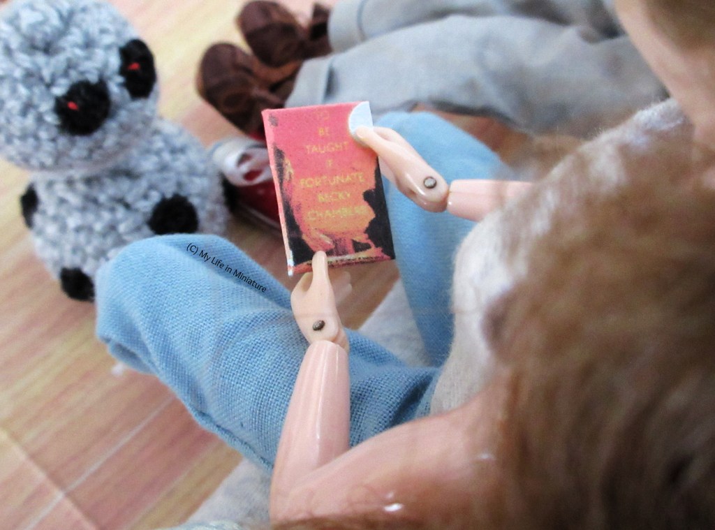 Over-the-shoulder shot of Sarah's hands, holding the orange book. We can see the cover now, which is that of 'To Be Taught If Fortunate' by Becky Chambers. Chip is in the background, looking on.