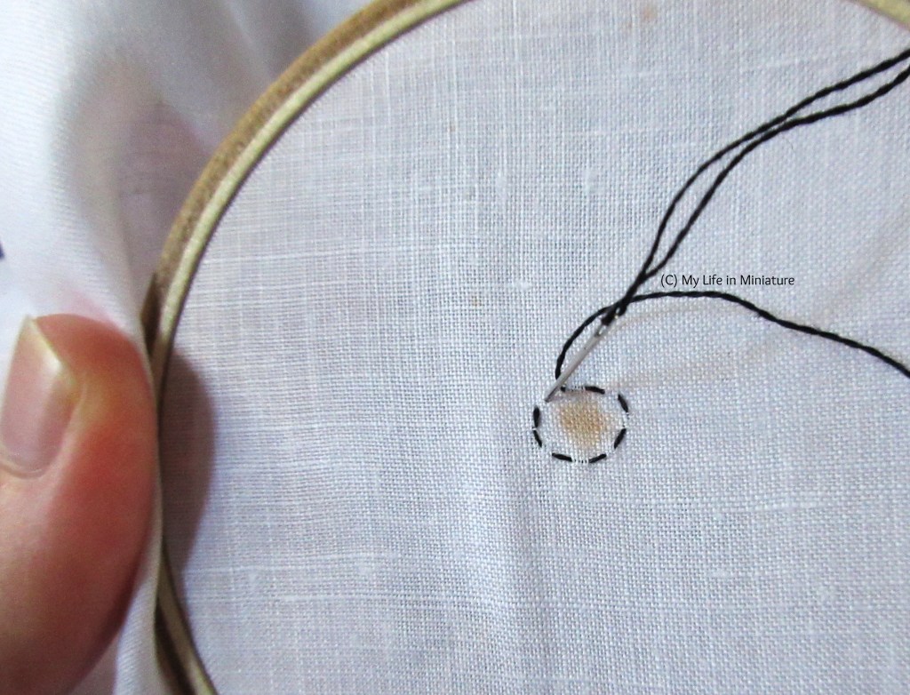 Close-up shot of one of the brown stains on the hankies. It's in an embroidery hoop and is being outlined in black thread. A hand is holding the hoop.