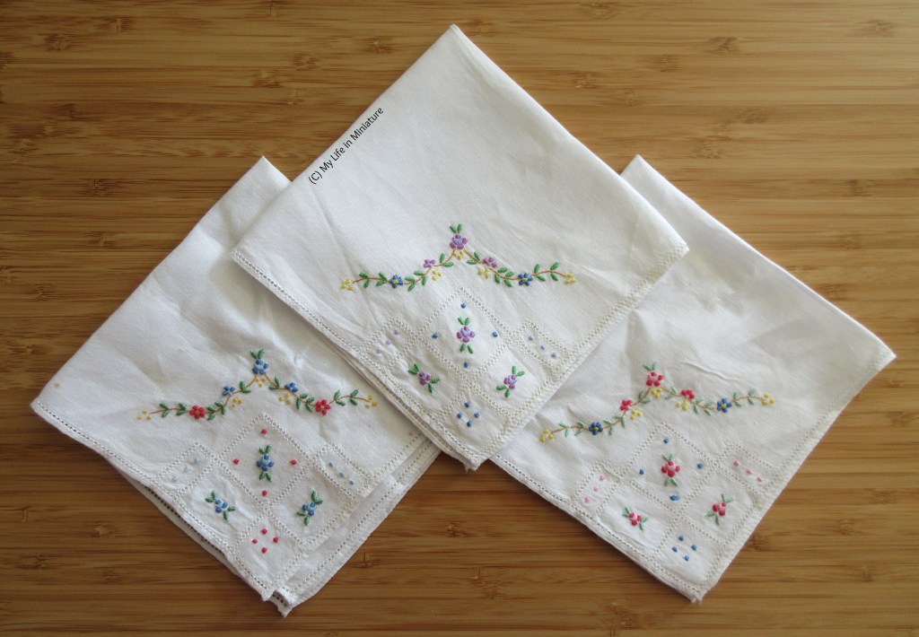 Three white handkerchiefs, folded into squares, sit flat on a wood background. The corners visible all have floral embroidery on them, as well as filet embroidery details. 