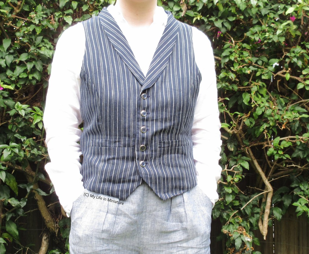 The author stands against a hedge. She wears a navy blue pinstriped waistcoat with a shawl collar and two single welt pockets. Under the waistcoat is a white linen shirt, and her hands are tucked into the pockets of grey linen pants. She faces the camera.