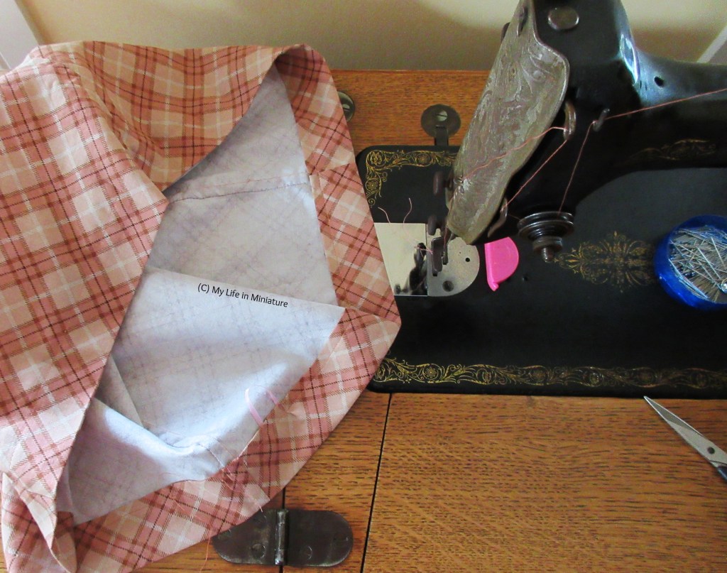 An overhead shot of the waistband of a pair of pants, sitting beside the needle-end of an old-fashioned sewing machine. The fabric is a peach and pink check, and the machine is threaded with similar coloured thread. Also visible is a pin tin and some embroidery scissors.