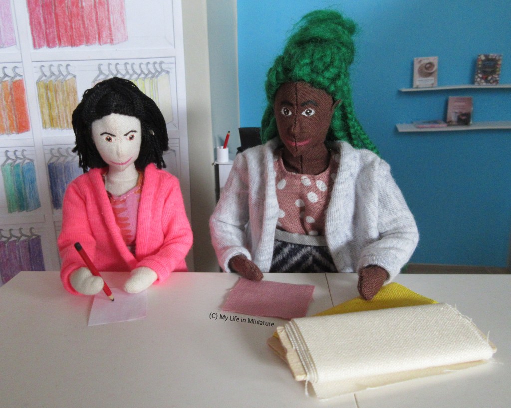 Hazel and Tiffany stand at the cutting table in Needle & Thread. Hazel has a sample square of dusky pink fabric and two bolts of fabric in front of her, and is describing something to Tiffany. Tiff is listening to Hazel, and has a pencil poised over a sheet of paper.