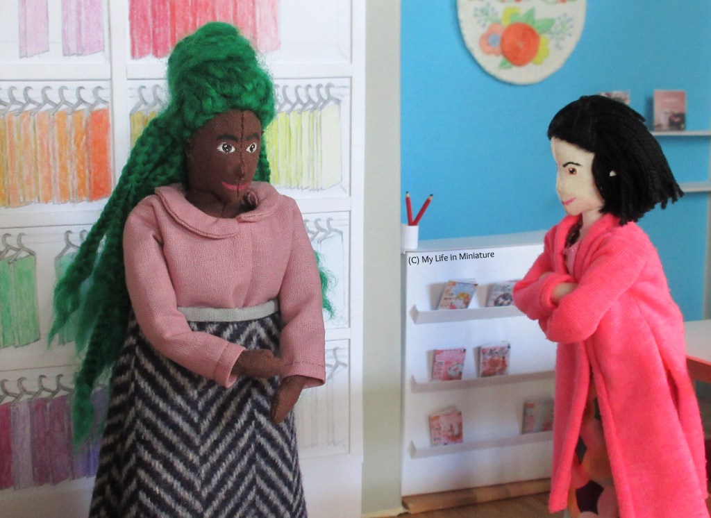 Hazel and Tiffany stand in Needle & Thread. Hazel has a dusky pink collared top on, with a long wool skirt. She is fiddling with the hem of one of the long sleeves. Tiffany watches on, arms crossed, wrapped up in her hot pink cardigan.