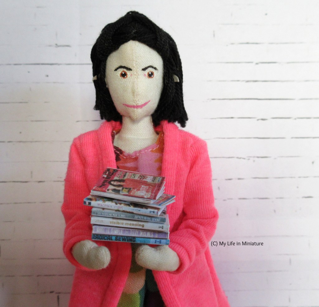 Tiffany stands in front of a white brick background. She holds a stack of craft books in her arms, with a couple of sewing magazines on top. 