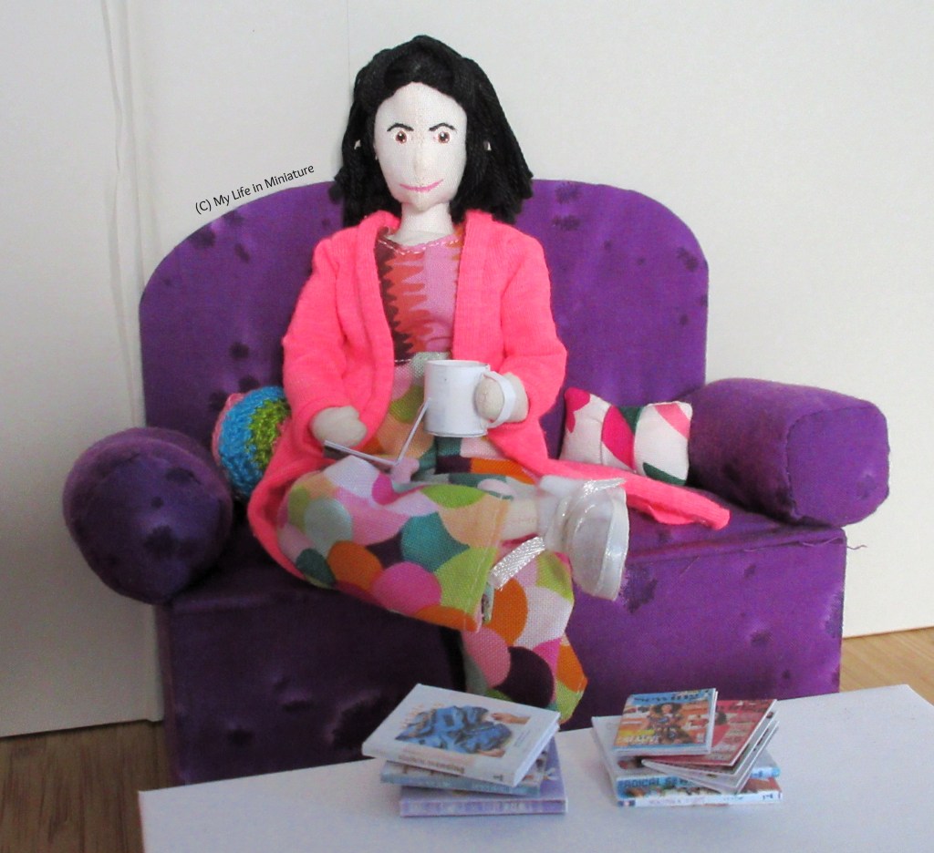 Tiffany sits on the purple couch in Needle & Thread's sit and stitch corner. She has an ankle resting on her knee, a cup of coffee in her hand, and a magazine open on her lap. On the coffee table in front of her are the other two magazines and the craft books, in stacks. She smiles at the camera. 