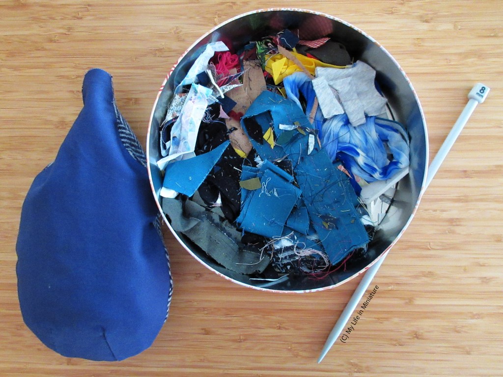 A partially-stuffed blue tailor's ham sits beside an open tin on a wood background. The tin contains various fabric and thread scraps. Beside the tin is a large knitting needle.