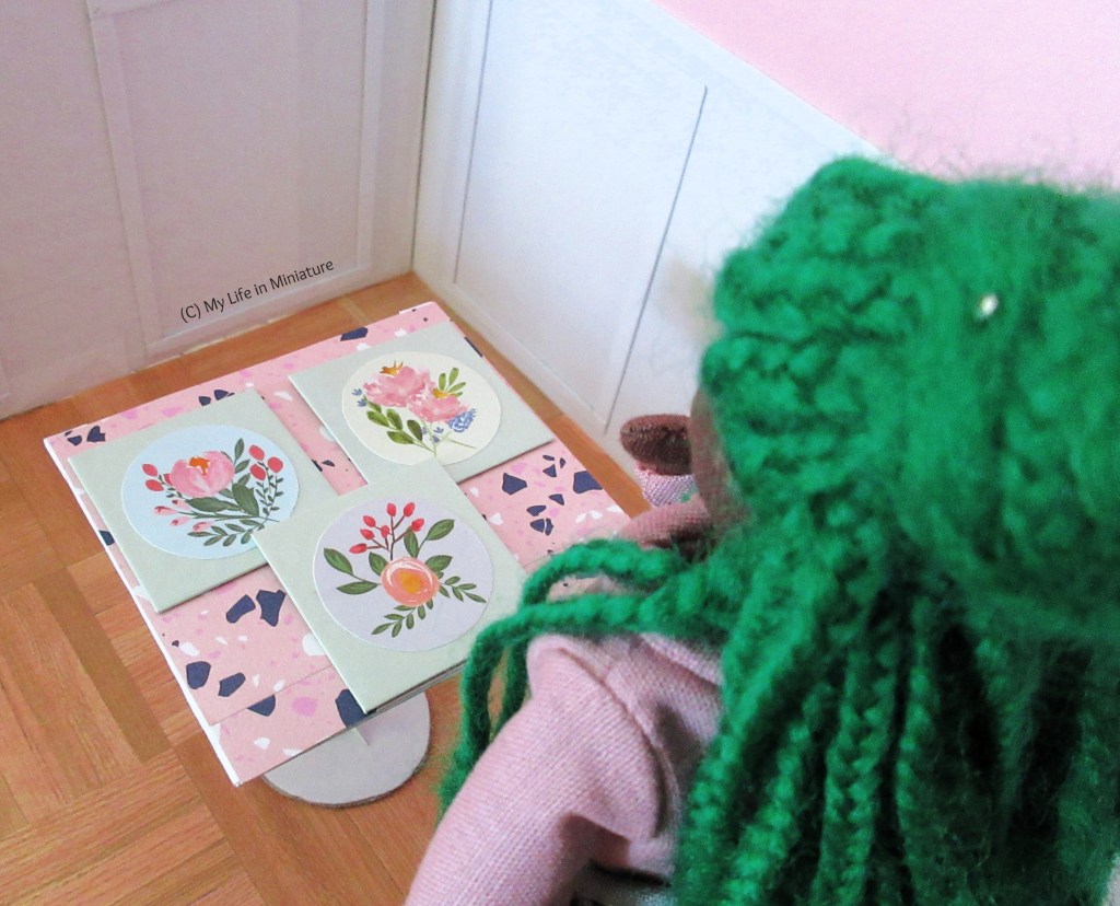 Hazel stands in Fierro's front room. The camera is looking over her shoulder at one of the marble-topped tables, which she's pulled into the corner. There are three square canvases on the table-top, and she has a hand to her chin in thought.