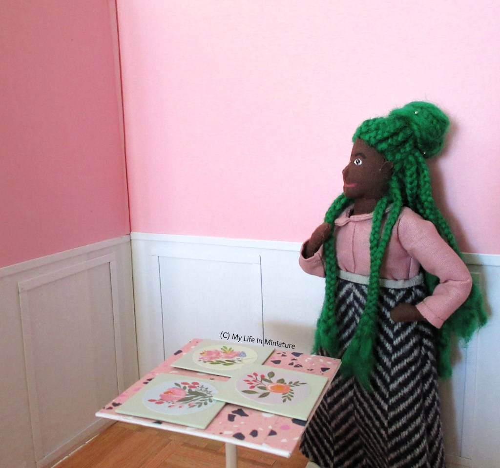 Hazel stands in the corner of Fierro's front room. The table is beside her, with the three canvases on it as before. She has a hand to her chin and the other on her hip as she looks up at the plain pink walls.