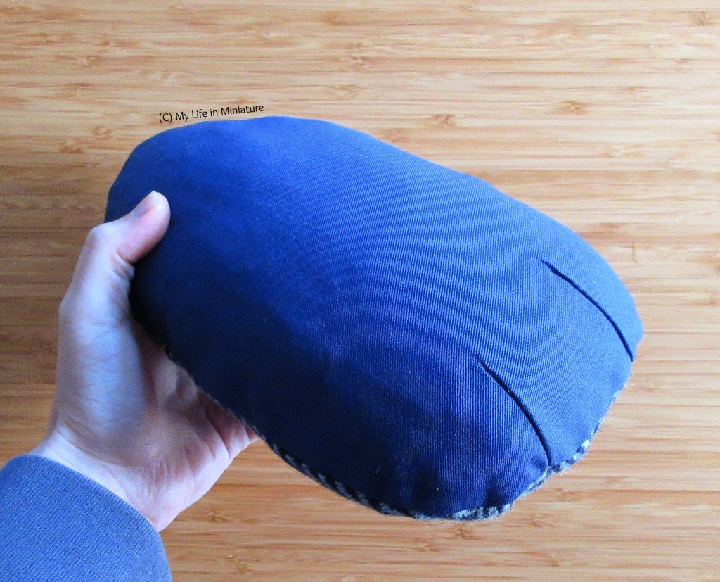 The author's hand holds a navy blue tailor's ham against a wood background. It is a vaguely egg-shaped cushion, stuffed very firmly. The rounder end is toward the camera, with two darts clearly visible.