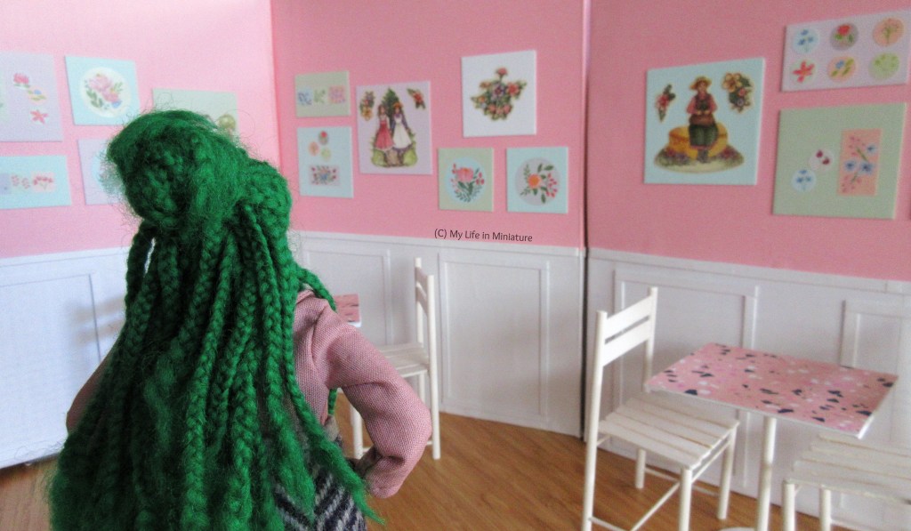 Hazel stands in the dining area of Fierro's Bakery. On the pink top halves of the walls, there are pastel canvases with floral art on them. Hazel stands in the foreground, back to the camera, hand on hip. Her green braids cover her back.