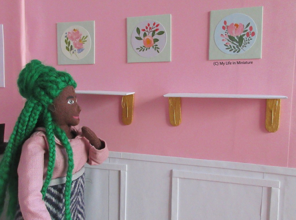 Hazel stands in front of the shelves, one hand raised to her mouth in thought. The trio of square canvases hangs above; they're all pale green with circular floral stickers in the centre.