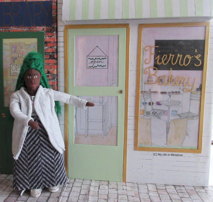Hazel stands outside Fierro's Bakery, gesturing towards it excitedly. Fierro's is clad in white brick, with a pale green door, gold frames, and gold writing on the window.