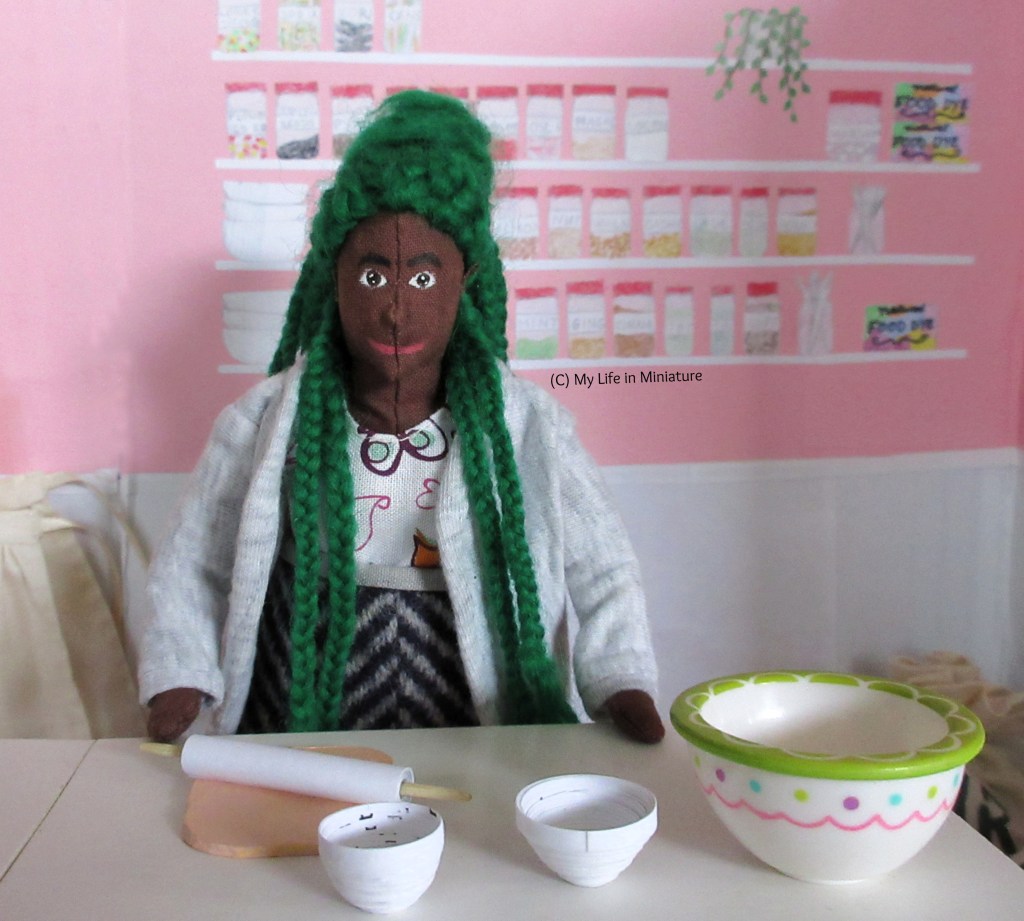 Hazel stands at the workbench, hands on the table. She smiles at the camera, a few of her braids falling forward over her shoulders. 