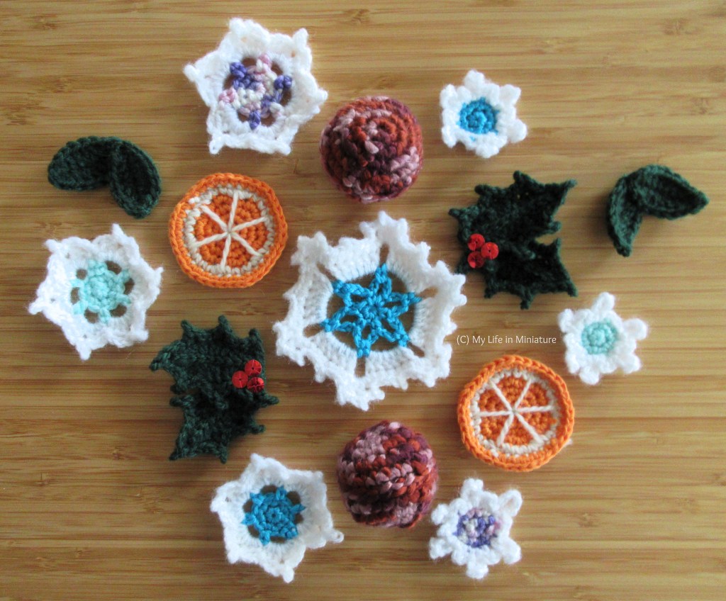 15 crochetd motifs sit, arranged in a loose group, on a wood background. The elements include about seven snowflakes of various sizes and colours, two pink baubles, two orange slices, and four pieces of foliage. Two of the foliage pieces are holly leaves with red sequins for berries. 