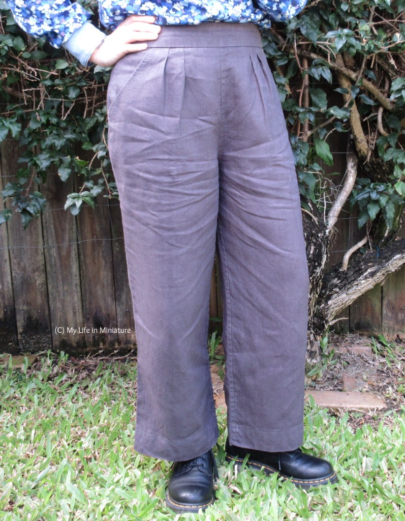 Another view of the grey pants worn by the author. Her other hand is on her hip, and her knee is slightly bent. There are two pleats on either side of the centre front, just near the pocket openings.