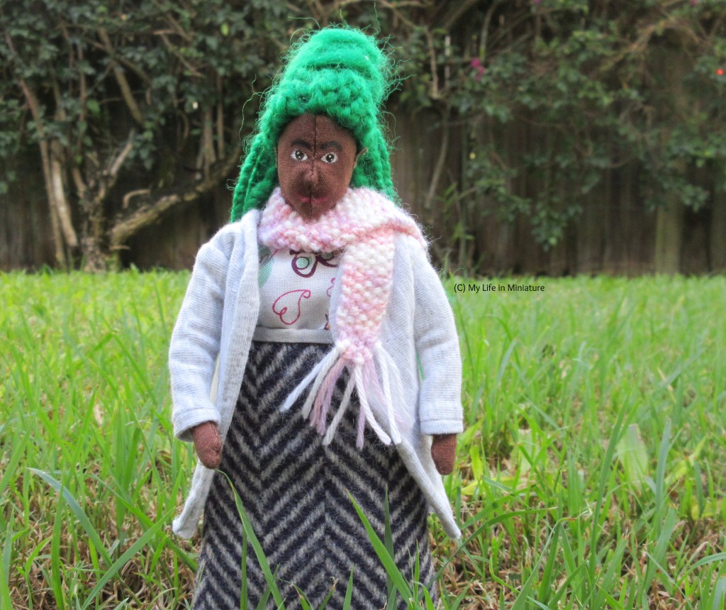 Hazel faces the camera, with grass and a fence behind her smiling face. She wears the pink and white vareigated scarf tossed over one shoulder, with a grey cardigan, wool skirt, and a shirt. Her arms hang at her sides. 