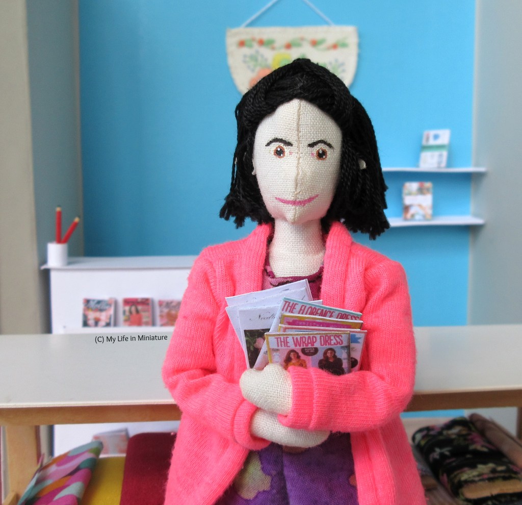 Tiffany stands in Needle & Thread, facing the camera and smiling. She holds a number of the sewing patterns in her arms.