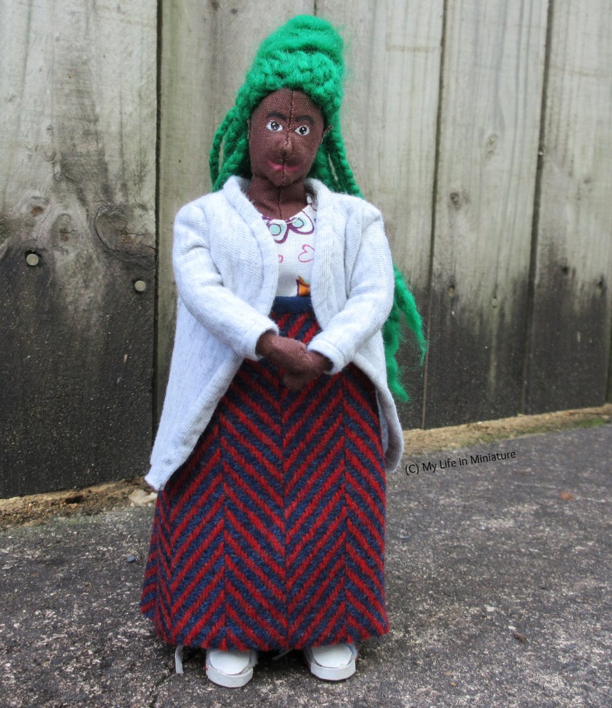 Hazel has her hands clasped in front of her as she smiles at the camera with her head slightly tilted. She's in her new wool skirt, with a grey cardigan and a white floral dress underneath.