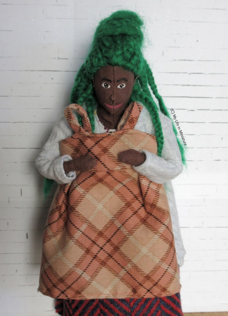 Hazel stands against a white brick background, holding a peach checked pinafore dress up to herself. She's in a grey cardigan and a wool skirt, and looks down at the dress.