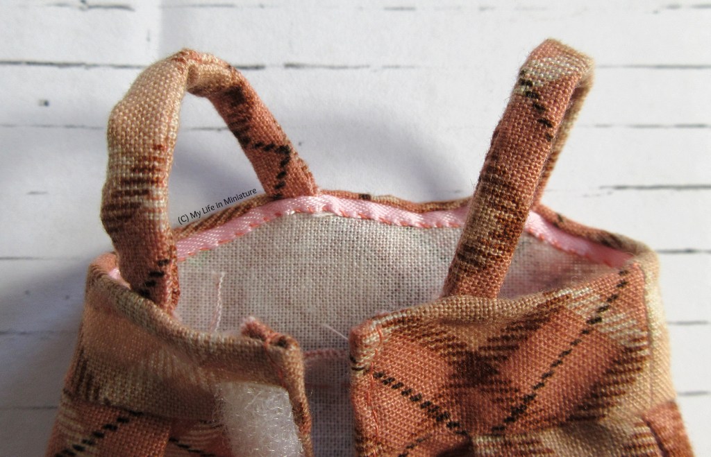Close-up of the interior of the top edge of the pinafore. A line of pale pink ribbon has been stitched over the hem edge, concealing the raw edges. The two straps match the fabric. 
