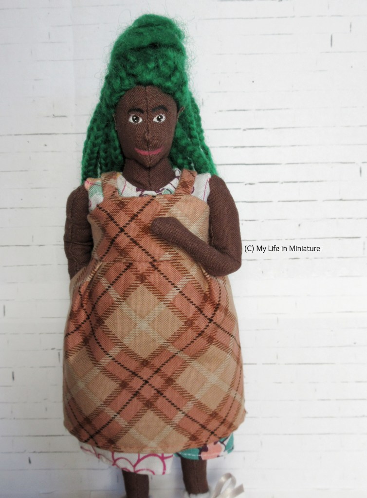 Hazel stands against a white brick background, smiling at the camera. She wears the peach pinafore over her white floral dress, one hand to her chest and the other in one of the in-seam pockets. 